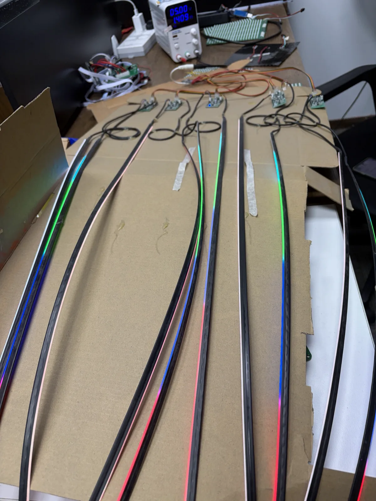 Ten fibre-optic ambient light strips laid out on cardboard on a workbench, each lit in a blue-green-pink gradient, each connected to its own small driver board and fed from a bench power supply.
