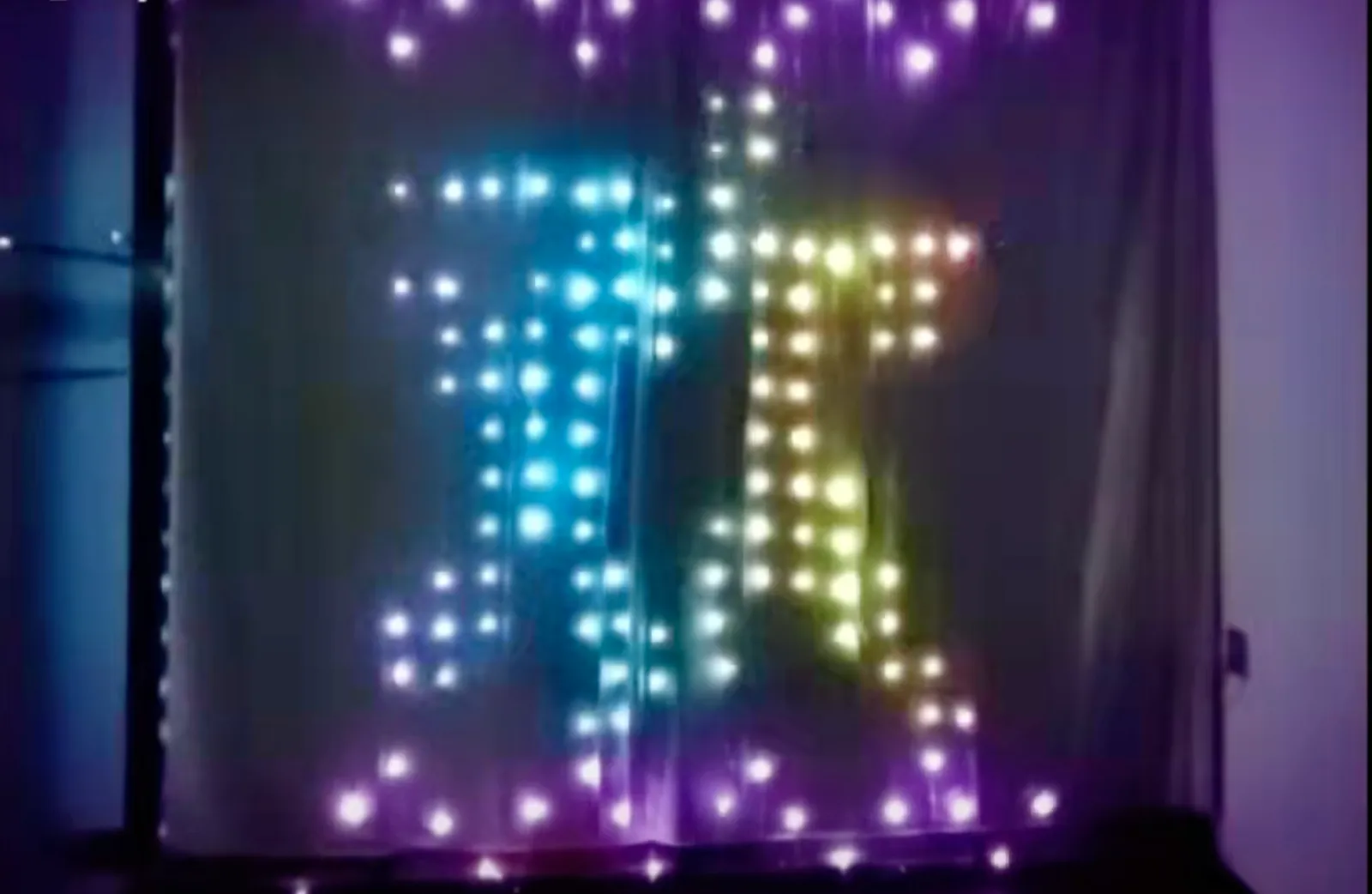 A pixel curtain hung across a window at night, its addressable LEDs forming a large glyph in cyan and rainbow colours against unlit black pixels.