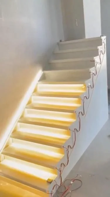 A white plastered staircase during construction, with warm white LED strip glowing under the nosing of the lower steps, the upper steps still dark, and loose red and black wires hanging down the open side of the flight, one pair to each step.