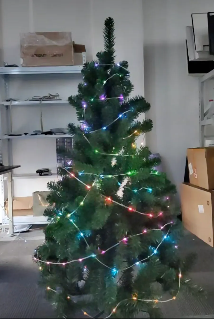 An artificial Christmas tree in a workshop with a single addressable LED string spiral-wrapped around it, the turns spaced far enough apart to leave visible gaps of unlit branches.
