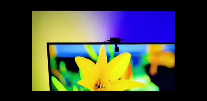 A desk monitor with a small colour-sensing camera stuck to the top edge and its cable running down, lit from behind by a soft red and cyan glow.